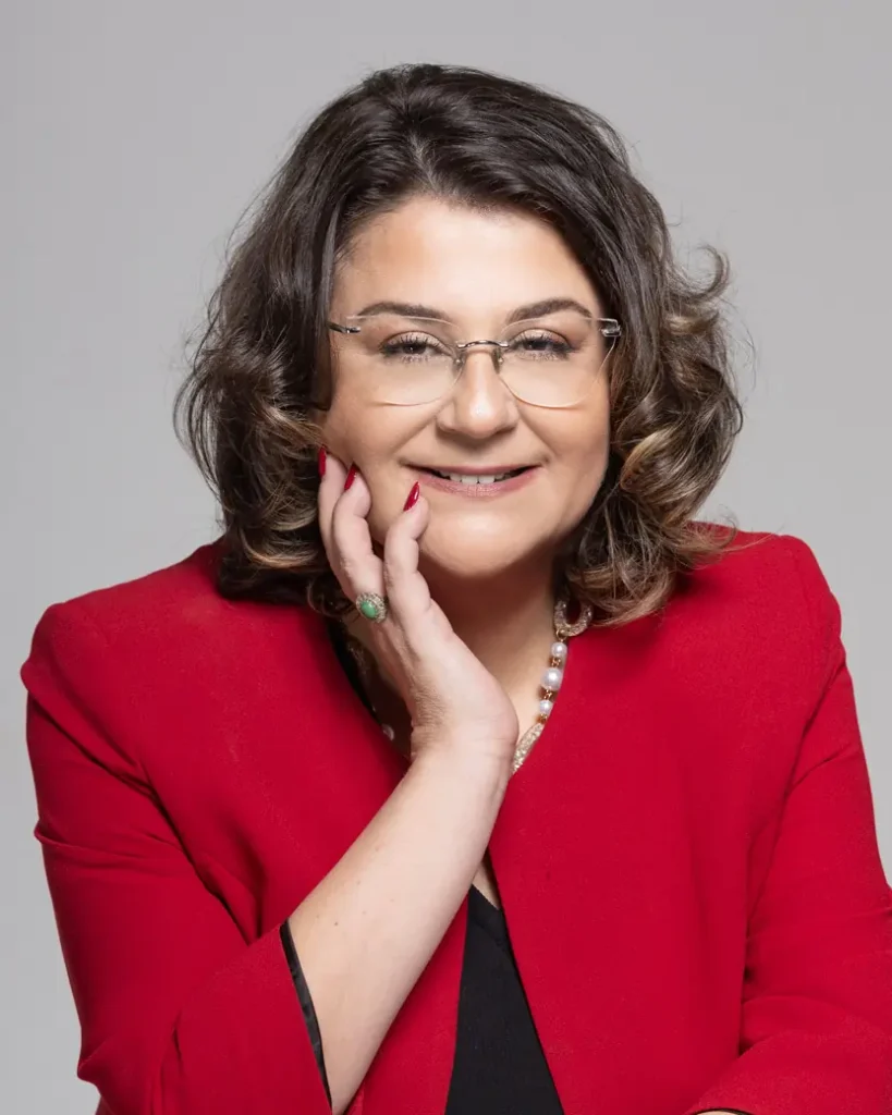 Dr. Menekşe Ahbab, kırmızı ceketle elini yanağına koymuş gülümserken
